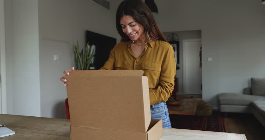 Woman stand at table opening large cardboard box with positive expression, enjoy received gift, got surprise or bought goods for home. Electronic commerce, webstore client, express transport services