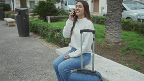 Woman talking on phone seated on park bench with suitcase beside wearing casual sweater and jeans in outdoor setting - Powered by Shutterstock - Get 15% off with code: PIKWIZARD15