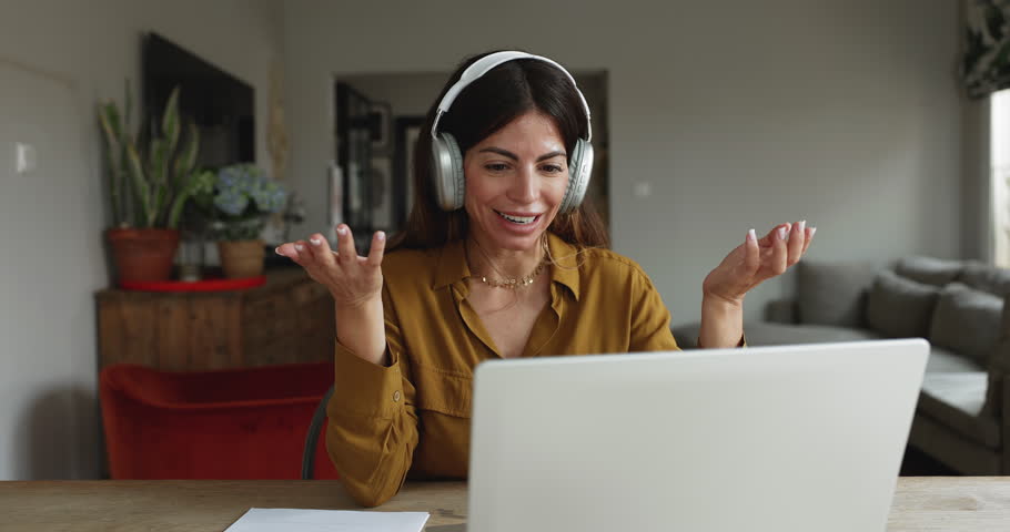 Woman in headphones sits at table with laptop conducting virtual meeting event with colleague or learner, teaching, leading briefing at home office using videocall app. Digital communication, tuition