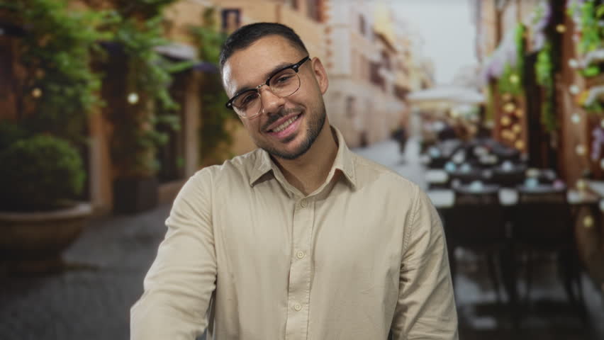Young hispanic man wearing glasses smiling with outstretched hand offering handshake on street; greeting friendliness.