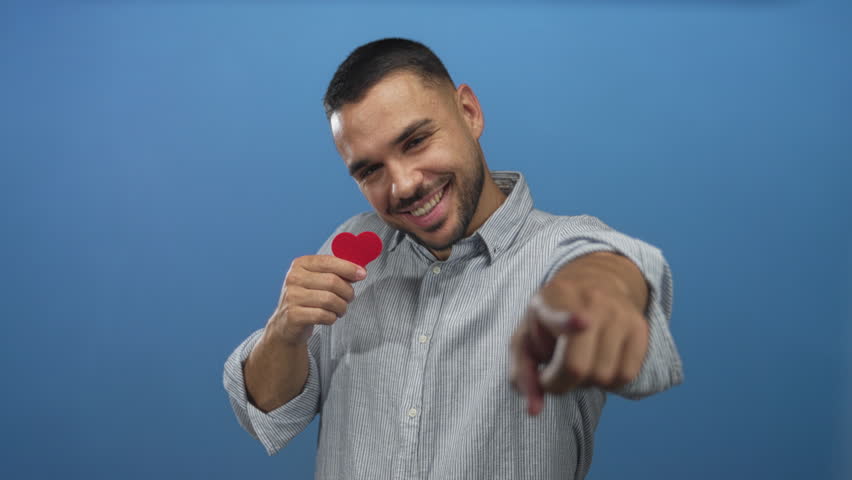 Hispanic man in striped shirt holding small red paper heart and pointing finger in blue studio; love connection.
