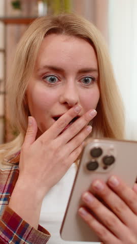 Young adult woman sits on home sofa reading surprising news on smartphone with wide eyes and curious look. Blonde girl reacts with awe wow and amazement to unexpected positive update or event online.