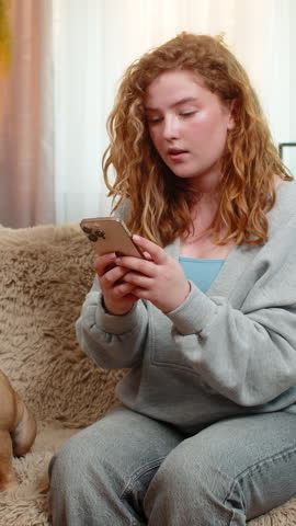 Young Caucasian redhead woman gets bad news on smartphone while sitting on home sofa, showing disappointment and tension. Girl frowns as pug dog nearby senses mood and reacts with curious posture.