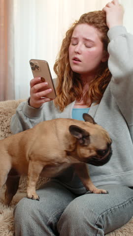 Young Caucasian redhead woman gets bad news on smartphone while sitting on home sofa, showing disappointment and tension. Girl frowns as pug dog nearby senses mood and reacts with curious posture.