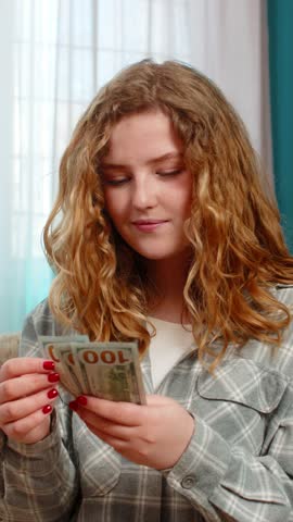 Young woman at home excitedly counting cash dollars in hands smiling and celebrating unexpected financial gain. Caucasian redhead girl on sofa happy about lottery win, salary or surprise money profit