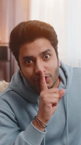 Shh be quiet please. Portrait of young Indian man presses index finger to lips makes silence gesture sign do not tells secret, stop talk gossip, confidential privacy. Guy sitting on sofa at home room.