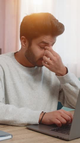 Young Indian man freelancer working on laptop, looking tired, rubbing eyes while sitting at table on sofa in living room at home. Arabian guy, a workaholic, continues his tasks, clearly exhausted.