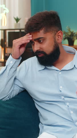 Indian man at home slaps forehead facepalm after smartphone error looks disappointed regretful frustrated. Arabian guy on sofa lowers head grimaces shows guilt mistake reaction stress negative mood.