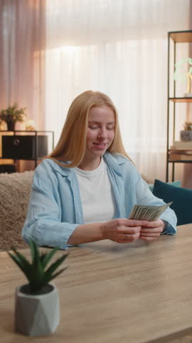 Young woman sits at home counting money dollar cash in hand, happy laughter from sudden lottery jackpot victory, salary, earnings. Caucasian girl waves banknotes celebrating financial surprise. Sunset