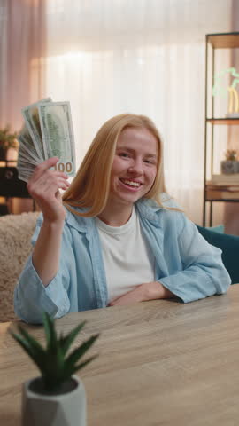 Young woman sits at home counting money dollar cash in hand, happy laughter from sudden lottery jackpot victory, salary, earnings. Caucasian girl waves banknotes celebrating financial surprise. Sunset