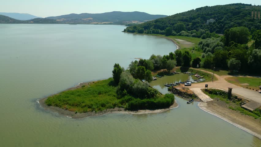 Aerial view of isola maggiore harbor, a small island on lake trasimeno, italy, showing boats moored near the shore, with green vegetation and hills in the background