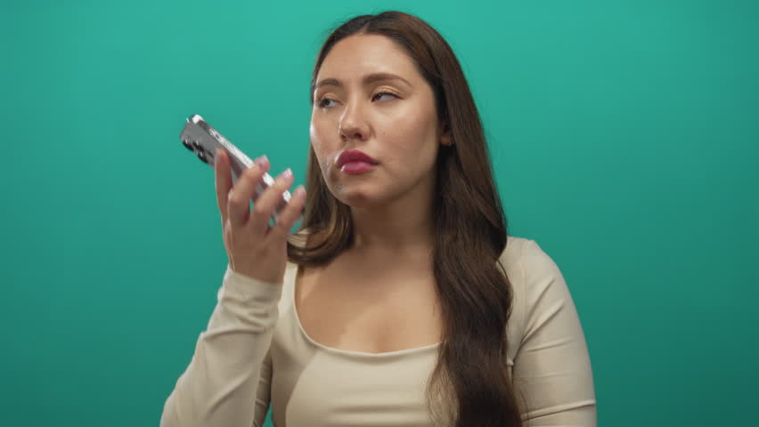 Young hispanic woman holds smartphone to ear and glances aside in studio with teal backdrop; focus.