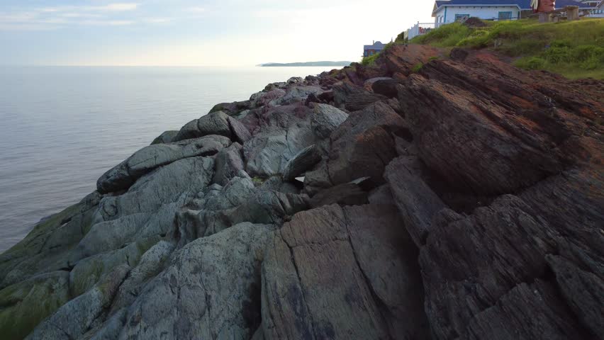 Scenic view featuring a rugged rocky coastline, calm ocean waters, and houses positioned on a grassy bank under a soft sky. Riviere-du-Loup, Quebec, Canada, 2025.