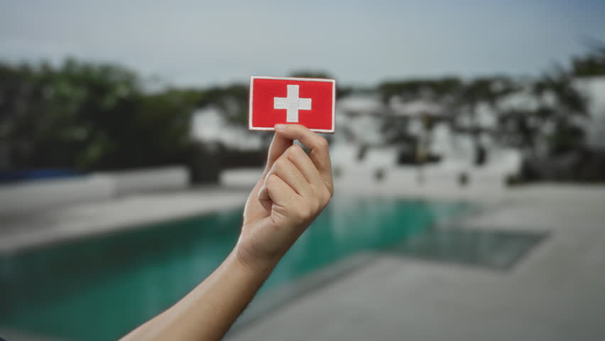 Man holding swiss flag patch above outdoor pool in a resort hotel setting, capturing a relaxed yet patriotic moment under clear skies.