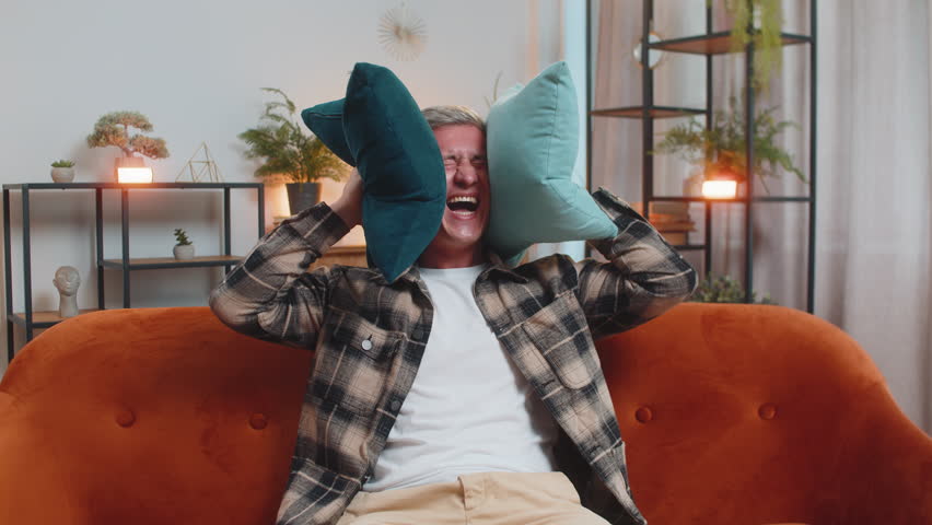 Repair work at neighbors. Irritated Caucasian man cover ears with pillows annoyed by noisy loud music neighbors suffer from headache wishes silence. Thin walls at home flat without sound insulation