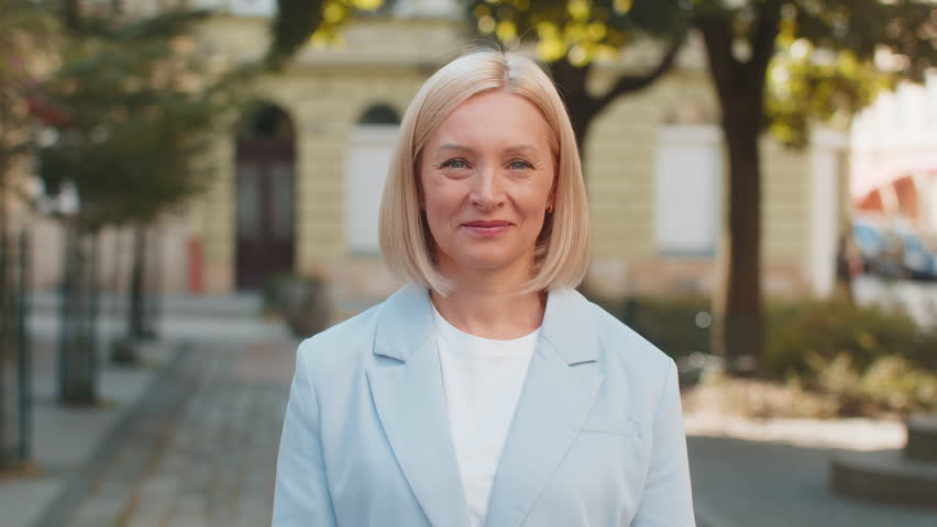 Happy smiling mature Caucasian blonde businesswoman lady female woman in formal suit looking camera and showing thumb up hand, positive sign gesture like recommend well-done outdoors city town street.