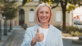 Happy smiling mature Caucasian blonde businesswoman lady female woman in formal suit looking camera and showing thumb up hand, positive sign gesture like recommend well-done outdoors city town street. - Powered by Shutterstock - Get 15% off with code: PIKWIZARD15