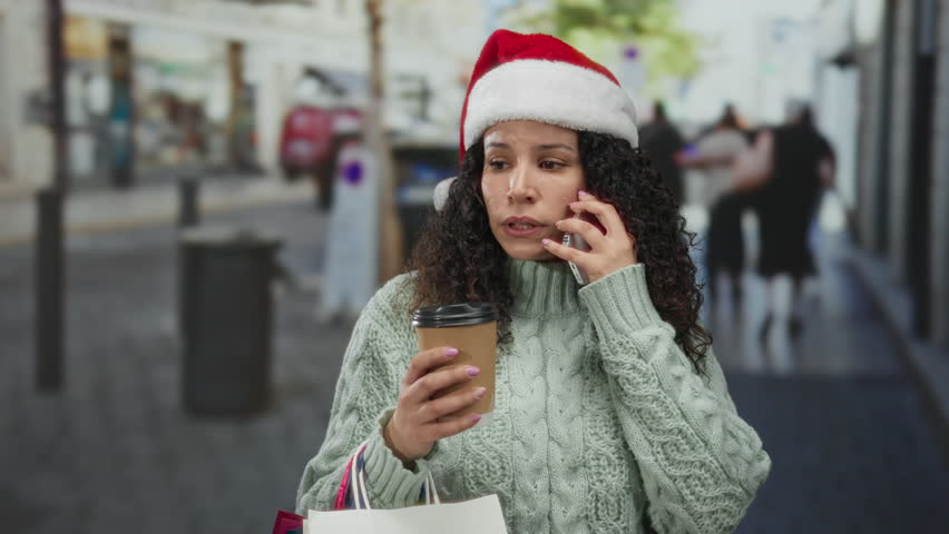 Woman wearing santa hat and sweater talks on phone holding coffee and shopping bags on a bustling city street.