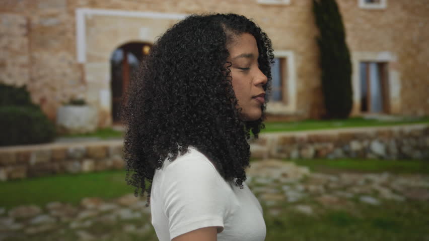 Woman displays curly hair, smiles warmly and wears a white t shirt at building entrance outdoors; confidence.