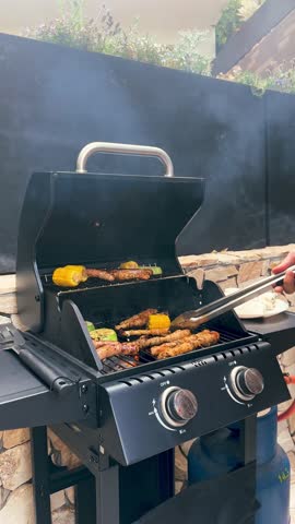 Person grilling meat skewers, sausages, and corn on a gas barbecue. Smoke rises from the grill while cooking outdoors. Tasty summer food preparation for picnic or backyard party.