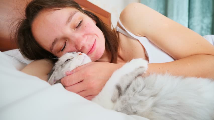 Young woman holds a kitten while sitting on a bed in a cozy bedroom, showing a tender moment between a domestic cat and its owner indoors.