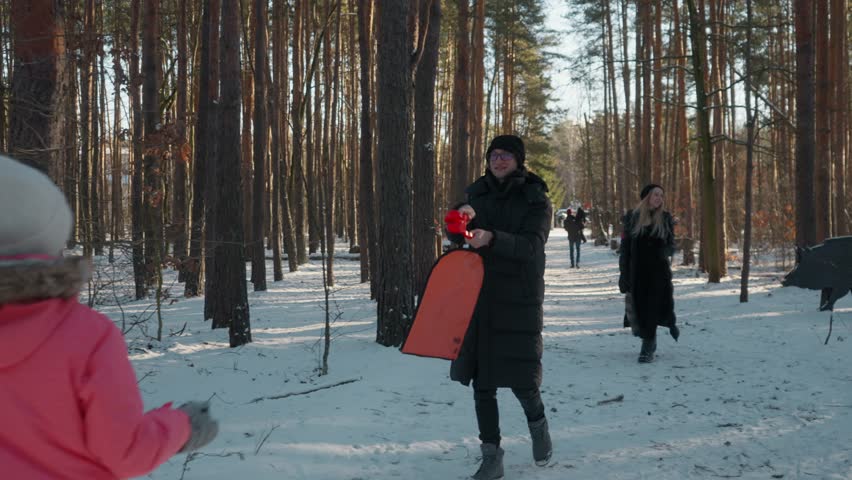 A young family with two children enjoys a winter walk through a snowy forest. Parents and kids carry sleds, play with snow, and spend active leisure time together outdoors on a beautiful sunny day.