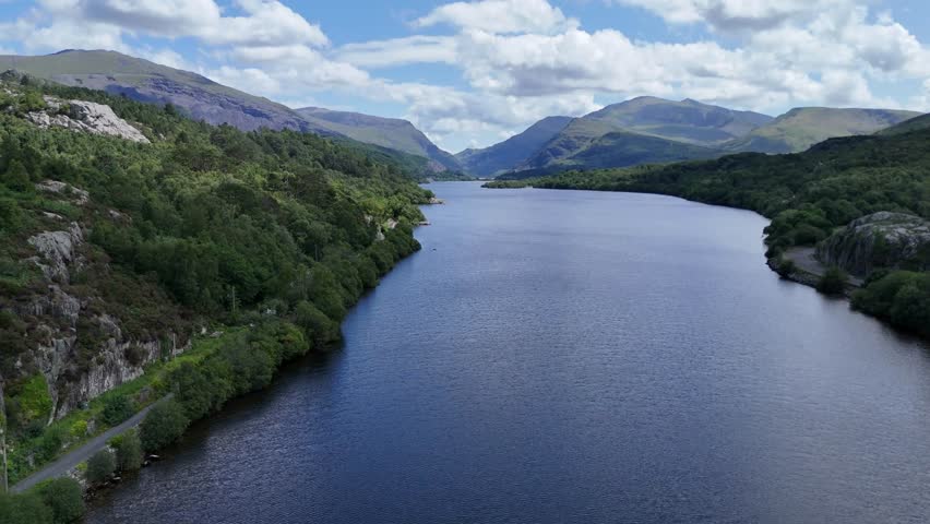Aerial drone footage flying over a serene river winding through a hilly landscape, surrounded by lush green trees and under a partly cloudy sky
