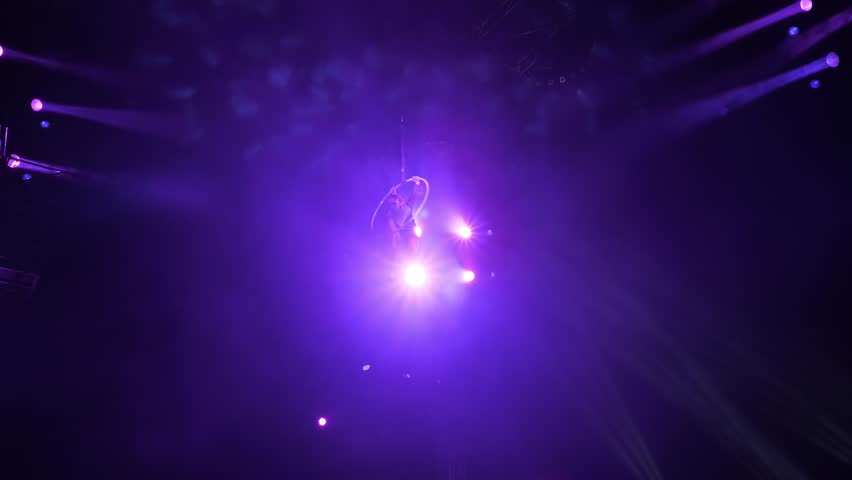 A slender aerial gymnast quickly rotates around herself on a gymnastic hoop under the dome of the circus. Aerial gymnast's performance, a cannon with smoke on the front panel. Circus show.