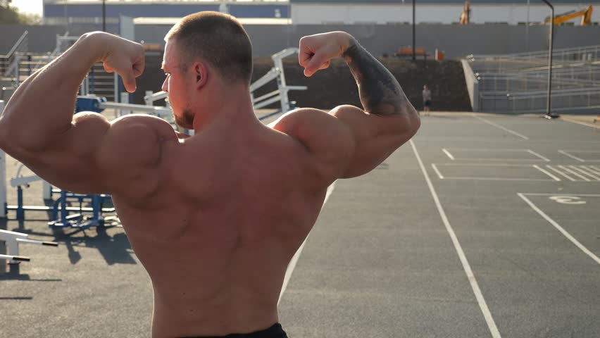 A handsome, muscular man shows off a toned back and arms. A bodybuilder shows off a toned body on an outdoor sports field at sunset. Outdoor exercise equipment. The image is stabilized.