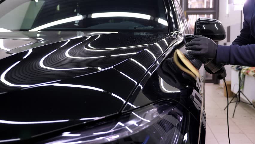 A close-up of a car mechanic polishing a black car with wax and a mechanical polishing machine. Modern car dealership and car body care.