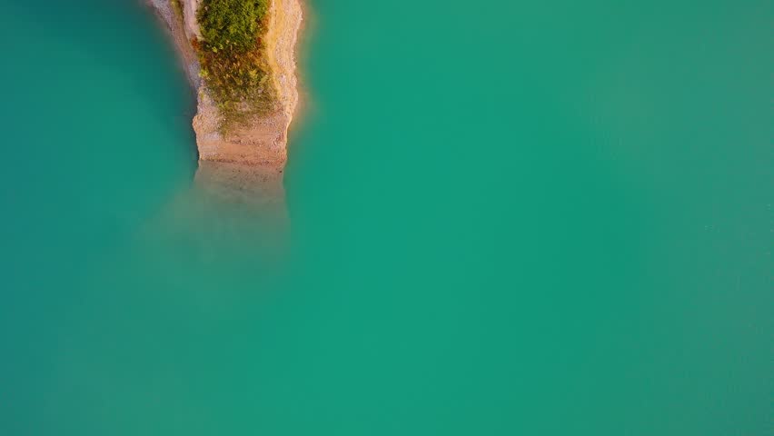 Drone video captures a rocky shoreline with green vegetation surrounded by bright turquoise waters of a lake in Spain, viewed directly from above.
