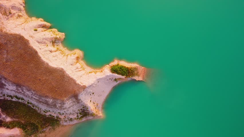 Drone video captures a rocky shoreline with green vegetation surrounded by bright turquoise waters of a lake in Spain, viewed directly from above.