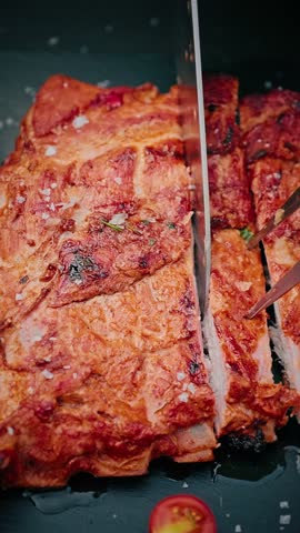 Close-up of juicy grilled pork ribs being sliced with a knife, sprinkled with coarse salt and herbs, served with cherry tomato on black slate.