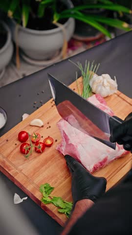 Cook wearing black gloves slices raw pork ribs on wooden board surrounded by cherry tomatoes, garlic, chives, peppercorns, and fresh herbs.