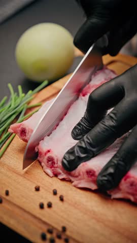 Close-up of chef wearing black gloves slicing raw pork ribs on a wooden board with onion, chives, and peppercorns nearby