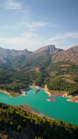 Drone video of the turquoise Guadalest Reservoir in Spain, surrounded by green hills and mountains, showing vivid water colors and scenic Mediterranean landscapes.