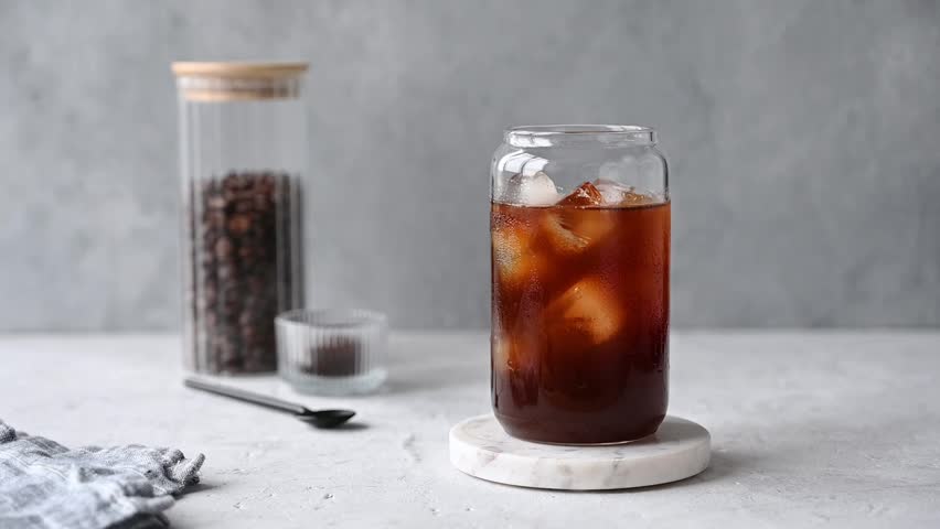 Milk pouring from a milk jug into a glass of iced coffee on a marble board on a light background with coffee beans. Trendy summer craft refreshing drink concept. Front view.