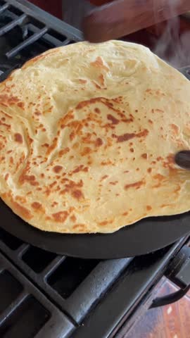 Buss up shot or paratha roti being cooked on a tawa (flat griddle or pan). A pair of dabla is also being used to handle the roti. This is being cooked in a kitchen in Trinidad, West Indies.