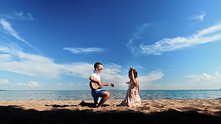 Two small children relax on sandy beach near lake on warm summer day: little boy plays guitar for beautiful girl against background of blue clear sky and water. Leisure time activity outdoors.