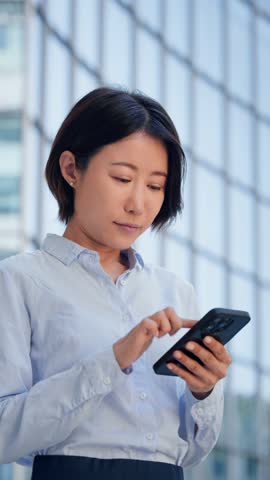 Focused busy female businesswoman holding smartphone for trading, banking, investing online outdoors on office background. Asian mature business woman manager ceo using cell phone mobile app. Vertical