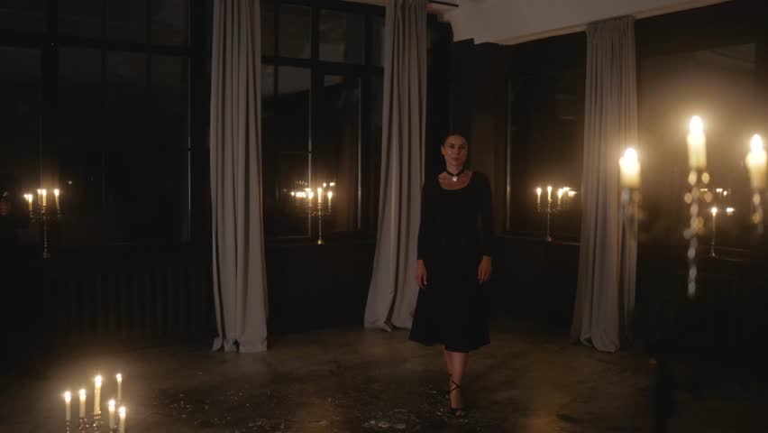 Mysterious woman wearing black dress and white heart-shaped necklace walking in dark room illuminated by multiple candles