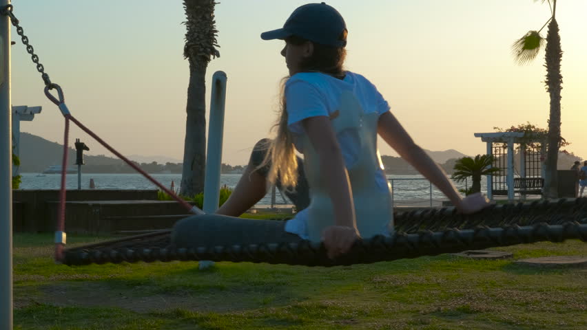 Girl gently rocking on a hammock by the sea at sunset. Relaxed girl gently swinging in rope hammock, basking in golden sunset light near coastal park with serene ocean horizon and grassy landscape