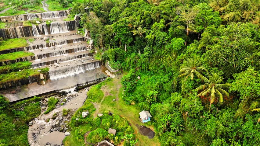 A stunning tiered waterfall cascading gracefully over stone steps, surrounded by lush tropical greenery and clear flowing water, located in Grojogan Watu Purbo Sleman Yogyakarta Indonesia.