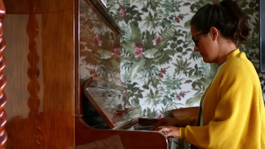 Skilled female pianist performing melodic composition on vintage brown upright piano, surrounded by ornate floral wallpaper in elegant interior setting