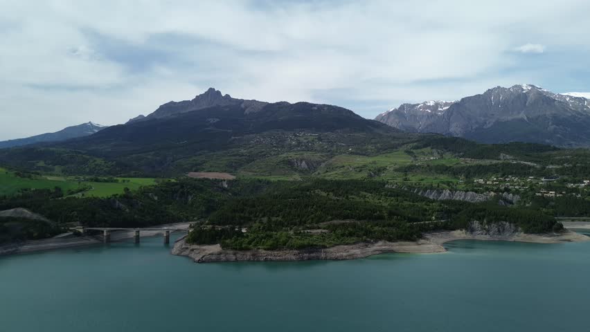 4K drone footage flying backward over Lac de Serre Ponçon, showcasing the Aiguilles de Chabrières, Parc des Écrins, a bridge, lush green trees, and snowy mountain tops. Captured May 2025, French Alps.