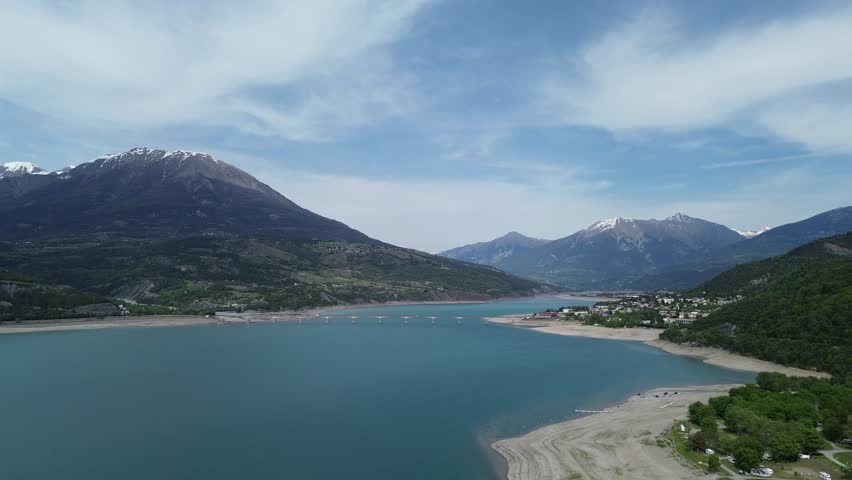 Crisp 4K drone footage of snowy mountains surrounding Lac de Serre Ponçon, focusing on Savines Le Lac Bridge and Mont Guillaume. Captured in the French Alps, May 2025, showcasing natural beauty.