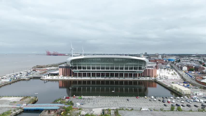 Modern architecture of the Hill Dickinson Stadium, reflecting on the calm waters of the dock, with wind turbines and cranes visible in the distance