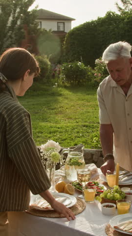 Vertical shot of mature man setting up dinner table in sunlit yard and meeting guests with embrace