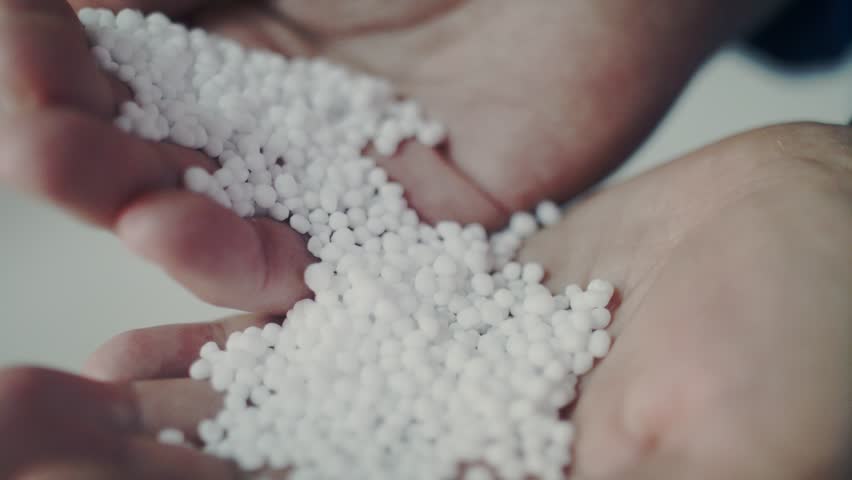 An extreme close-up shot of a person's hands inspecting white, spherical fertilizer granules. The pellets are poured from one hand to another, some falling in slow motion