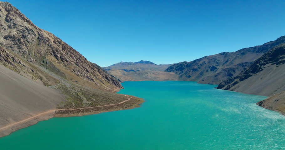 Drone flight over turquoise water of Embalse El Yeso reservoir in Cajon del Maipo, rugged Andes mountains and dirt road under clear blue sky, Chile. Aerial forward, copy space